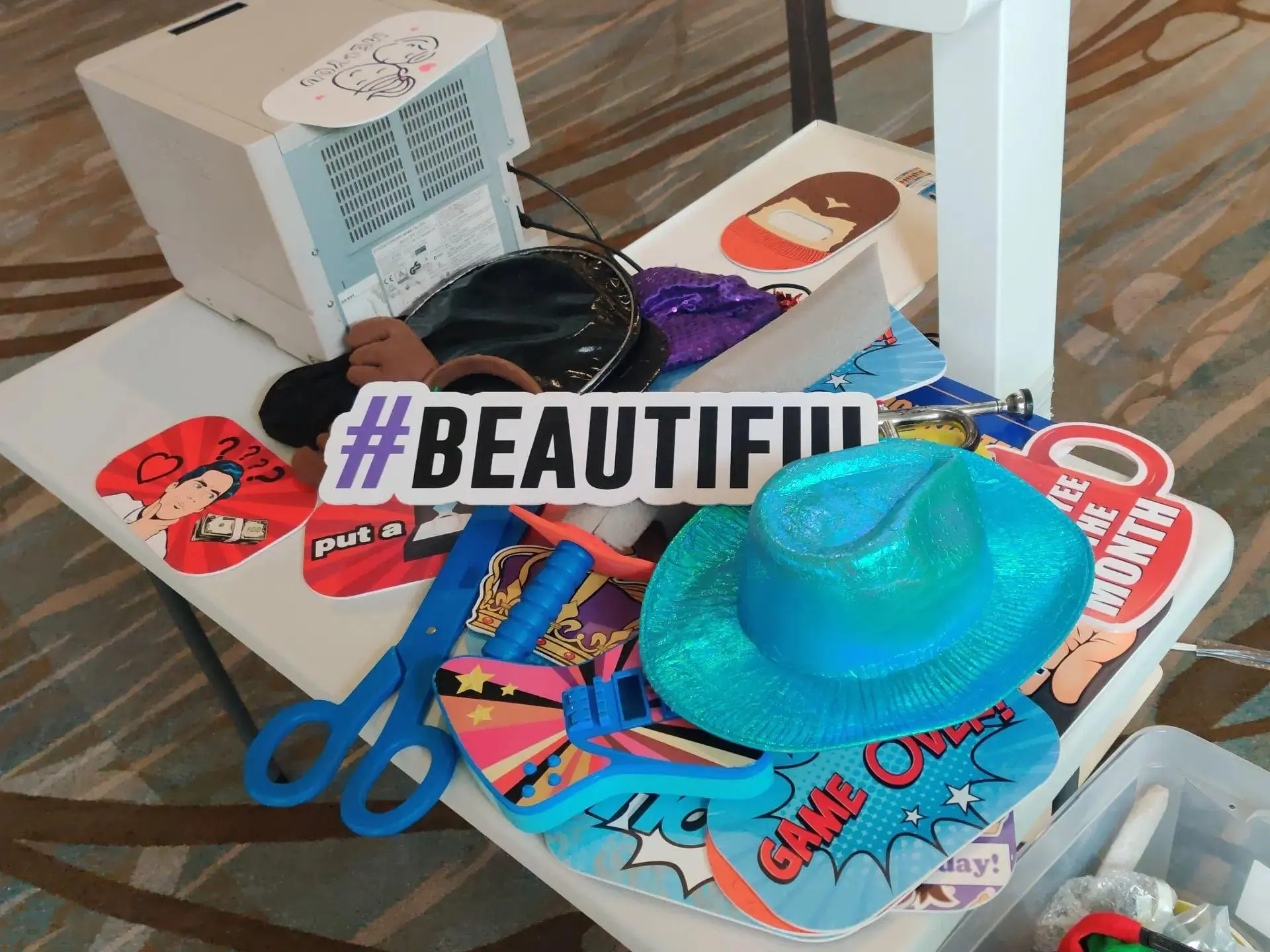 Colorful assortment of photobooth props, including a blue glitter hat, oversized sunglasses, and playful signs with hashtags and phrases, laid out on a table beside a photobooth printer, ready for guests to enjoy at Rent a PhotoBooth photobooth rental events.