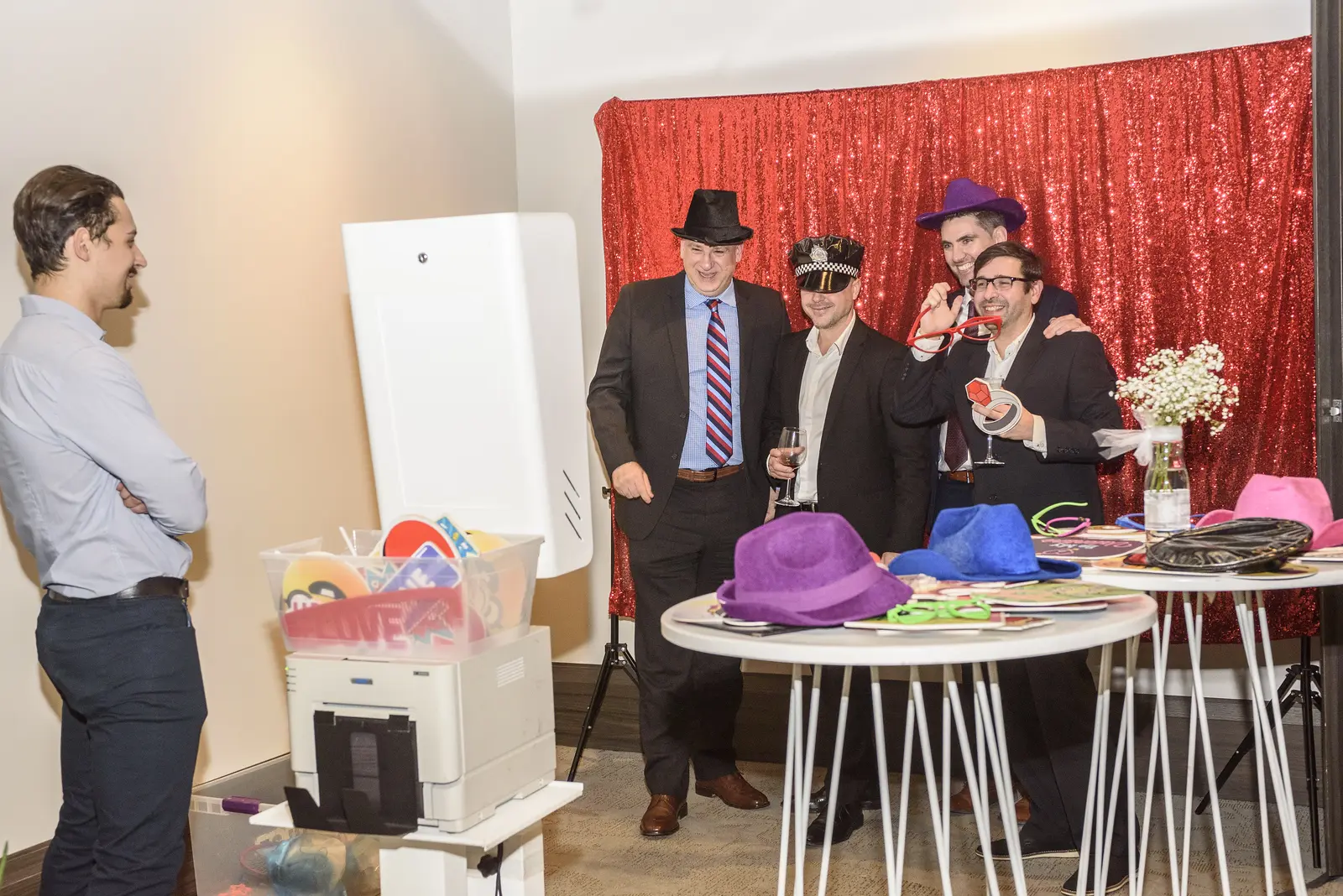 Corporate event attendees enjoying a photobooth with a vibrant red sequin backdrop, trying on various fun hats and glasses from a prop table, with a photobooth kiosk and printer in the scene, provided by Rent a PhotoBooth photobooth rentals.