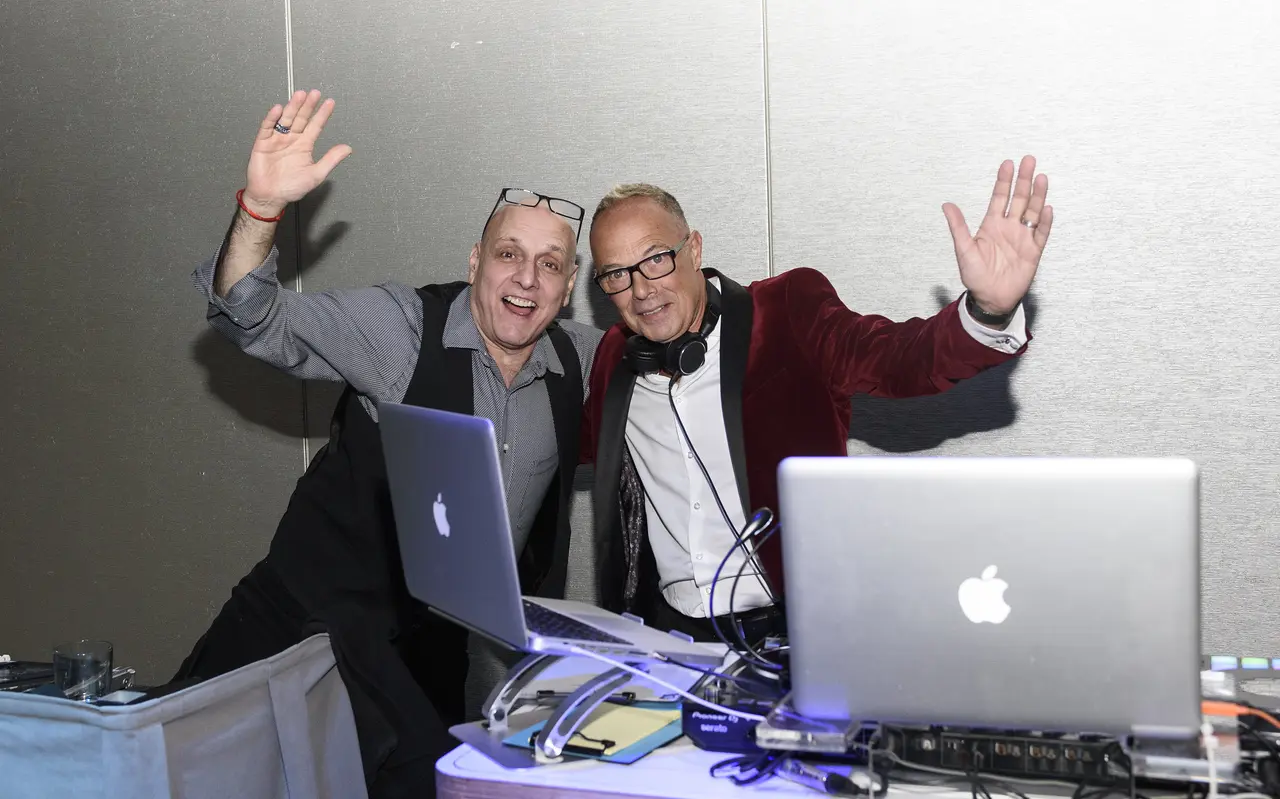 Shot by our professional event photographer, this image captures two exuberant DJs enjoying the party, with one wearing a stylish red velvet jacket and the other in a smart vest, both waving to the camera behind a setup of Apple laptops and DJ equipment, exemplifying the dynamic entertainment and high-quality photography services we offer for special events.